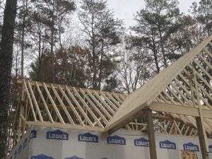 Cabin construction in progress, showing exposed wooden framing and roof rafters under a cloudy sky. "Lowe's" branded weather barrier visible on the cabin's exterior walls. Build your dream cabin with our company!