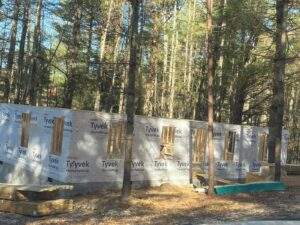 A half-built home stands framed by tall trees, its Tyvek HomeWrap exterior contrasting with the wooded backdrop. Lumber stacks hint at ongoing construction, inviting viewers to build with our cabin building company.