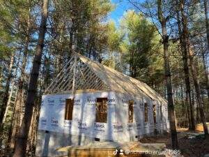 A cabin under construction in a wooded area, showcasing the framing and Tyvek wrap. Sunlight filters through the trees, highlighting the building process of this custom-built home by our cabin building company.