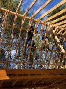 Two construction workers build a cabin frame in the Red River Gorge, Kentucky. The wooden beams form a geometric pattern against a blue sky and trees. "Boise Cascade Versa-Lam LVL" is visible on the lumber.