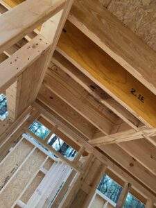 Cabin construction detail: exposed framing shows studs, beams labeled "2.1E," and OSB sheathing. Sunlight streams through window openings, highlighting the building process of a custom home in Kentucky.