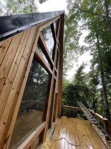 A-frame cabin construction in Red River Gorge, Kentucky. Large windows reflect the surrounding forest, showcasing the cabin's modern design and natural setting. A ladder leans against the deck, hinting at ongoing work. Build your dream cabin with us!