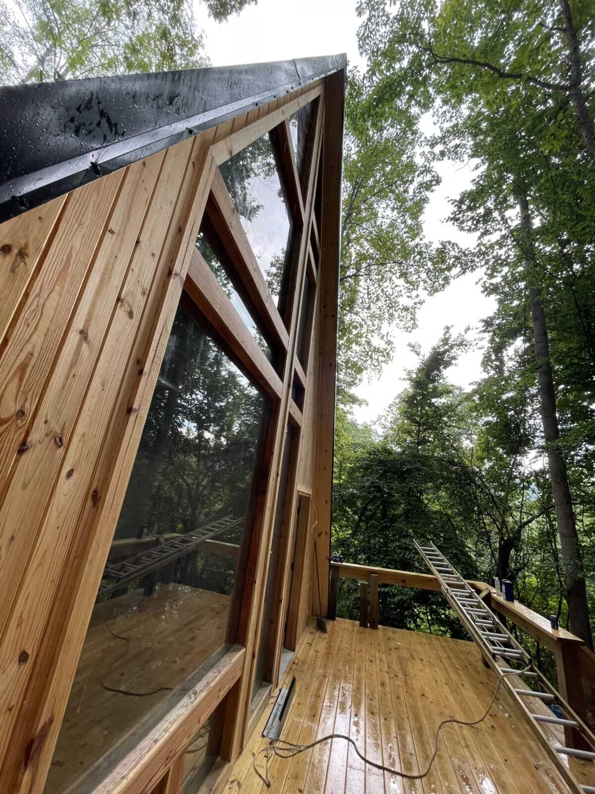 A-frame cabin construction in Red River Gorge, Kentucky. Large windows reflect the surrounding forest, showcasing the cabin's modern design and natural setting. A ladder leans against the deck, hinting at ongoing work. Build your dream cabin with us!