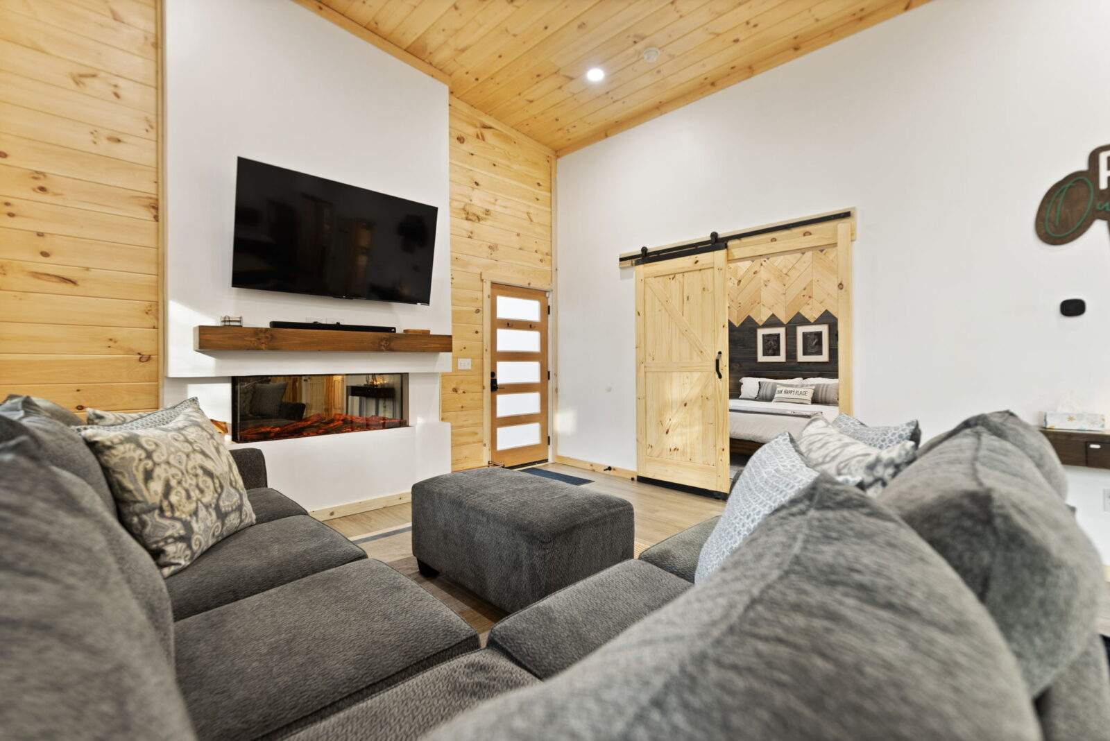 Cabin interior featuring a gray sectional sofa, modern fireplace, and sliding barn door leading to a bedroom. The wood paneled ceiling adds warmth, highlighting the luxury cabin's design for Airbnb investors.